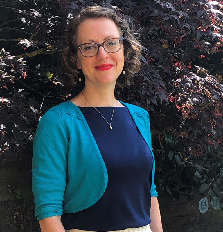 Photo of a white woman dressed in blue in front of a dark purple tree. She has red lipstick, glasses and a friendly smile